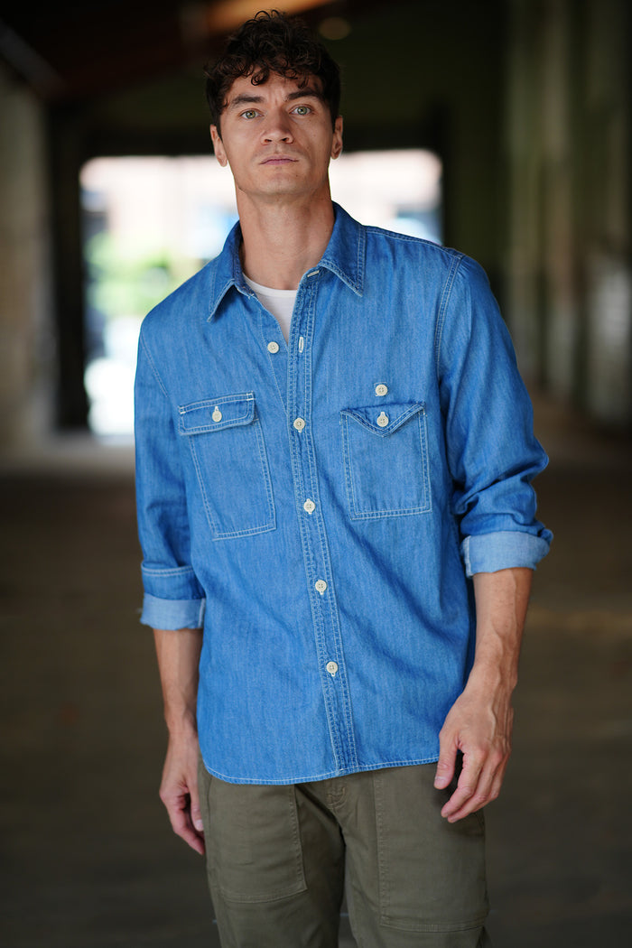 Man wearing a blue chambray shirt standing in a hallway setting.